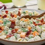 A candid, naturally lit photo of a large rustic serving bowl filled with creamy bacon ranch pasta salad loaded with cheddar cheese, crispy bacon, and peas on a wooden picnic table.