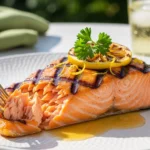A close-up of flaky, perfectly cooked Grilled Honey Lemon Butter Salmon topped with fresh parsley and a grilled lemon wheel on a white plate.