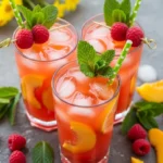 A large glass pitcher filled with bright pink homemade raspberry peach lemonade, fresh peach slices, whole raspberries, and mint leaves on a sunlit wooden patio table.