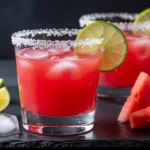 A candid, human-shot photo of a bright pink fresh watermelon margarita recipe served over ice with a salted Tajín rim, sitting on a sunny wooden patio table.