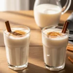 A naturally lit, candid photo of a woman's hand pouring a creamy, authentic horchata recipe from a glass pitcher into an ice-filled glass garnished with a fresh cinnamon stick.