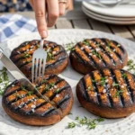 A woman's hands using a knife and fork to slice into a juicy, cross-hatched grilled portobello mushroom topped with fresh thyme and sea salt on a white platter at a sunny outdoor BBQ.