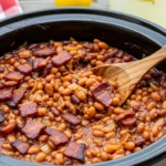 A candid, naturally lit photo looking down at a rustic black crockpot filled with thick, sticky slow cooker bourbon baked beans heavily garnished with crispy bacon on a backyard picnic table.