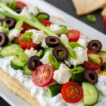 A stunning long white platter serving an easy Mediterranean appetizer of 7-layer Greek dip, featuring a smooth hummus and cream cheese dip topped with fresh cucumbers, cherry tomatoes, sliced olives, and feta cheese.