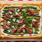 An overhead top-down photo of a whole golden-brown asparagus puff pastry tart resting on a wooden cutting board, showcasing its flaky crust and topped with roasted asparagus and prosciutto.