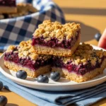 An extreme close-up photo of two Blueberry Oatmeal Crumble Bars, straight-on view to emphasize the distinct internal layers of sturdy oat base, glistening fruit filling, and crunchy crumble topping.