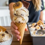 A candid photo of a person holding a waffle cone stacked with two scoops of no churn s'mores ice cream, featuring a toasted marshmallow and chocolate bar, with a loaded metal loaf pan in the background.