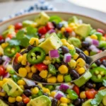 An appetizing, extreme close-up of a silver spoon lifting a generous, loaded scoop of the best Cowboy Caviar ever right out of the bowl, featuring glistening black beans, yellow corn, a sliced jalapeño ring, and chunks of creamy avocado.