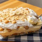 A warm, inviting photograph of the best easy banana pudding served in a glass dish, beautifully topped with fluffy whipped cream, crushed cookies, and fresh banana slices on a sunlit wooden table.