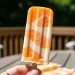 A candid, natural photo of a hand holding a swirled orange creamsicle dessert on a sunny outdoor patio, showing the creamy texture and frosty condensation.