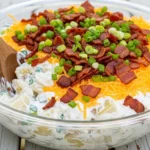 A large glass serving bowl filled with creamy loaded baked potato salad, topped generously with crispy bacon, shredded sharp cheddar cheese, and fresh green onions, resting on a white wooden picnic table.