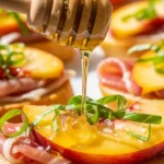 A mouth-watering close-up of a wooden honey dipper drizzling sweet honey over a grilled peach crostini with honey, fresh basil, flaky sea salt, and sliced prosciutto.