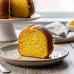 A beautifully baked 1920 Famous Ritz Carlton lemon pound cake, lightly dusted with powdered sugar and resting on a dark slate board. Fresh lemons and a microplane zester sit in the soft, brightly lit background.