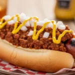 A close-up, sunlit photo of two grilled hot dogs in soft buns, generously topped with savory slow cooker hot dog chili, diced white onions, and bright yellow mustard on a clean white plate.
