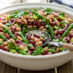 A candid, bright photo of a large white bowl overflowing with a classic three bean salad recipe, featuring fresh green beans, white beans, and kidney beans on a rustic wooden picnic table.