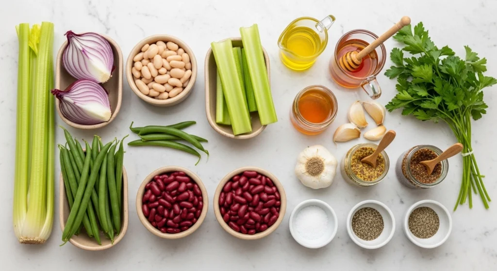 A bright overhead flat lay on a white marble counter displaying the fresh ingredients for a three bean salad, including celery, red onions, three types of beans, olive oil, honey, garlic, and fresh parsley.
