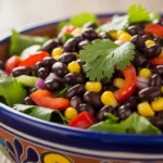 A close-up view of an authentic Mexican black bean salad served in a decorative ceramic bowl, packed with sweet corn, juicy tomatoes, red bell peppers, and topped with a fresh cilantro sprig.