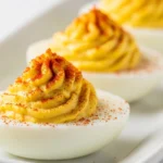 A close-up of classic deviled eggs lined up on a white plate, showcasing a perfectly smooth, creamy piped yolk filling dusted with bright red paprika.