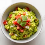 A clean, top-down view showing how to make guacamole from scratch, served in a minimalist ceramic bowl on a marble countertop and garnished with colorful diced tomatoes.