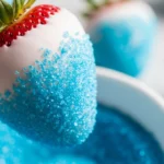 A bright, appetizing photograph of a white plate filled with gorgeous patriotic fruit desserts, featuring fresh red strawberries dipped in creamy white chocolate and bright blue sugar, sitting next to a festive American flag napkin.