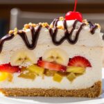 A direct overhead shot of a fully assembled No Bake Banana Split Dessert in a rectangular glass baking dish, topped with fluffy whipped cream, a beautiful lattice of chocolate syrup, chopped walnuts, and bright red maraschino cherries.