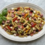 A direct overhead shot of a colorful, fully assembled antipasto pasta salad served on a decorative white oval platter. The salad is packed with rotini pasta, thick-cut salami, pepperoni, provolone cheese, artichoke hearts, roasted red peppers, and olives, garnished with a sprig of fresh basil.
