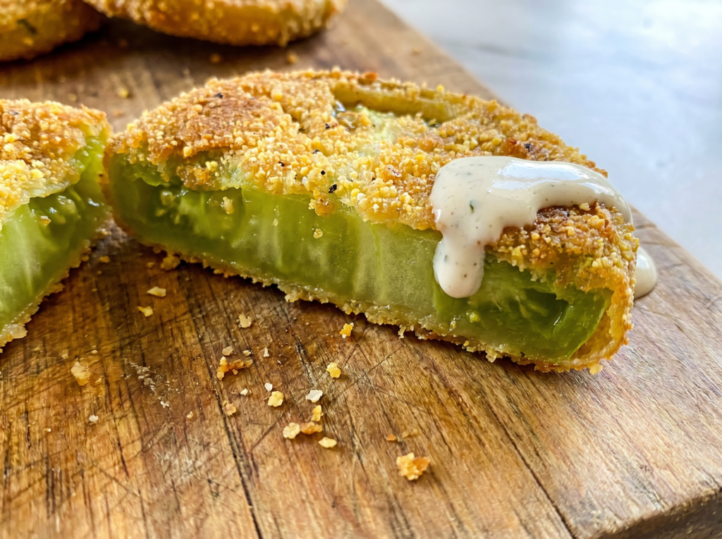 A macro cross-section of a fried green tomato on a wooden board, showing the contrast between the tart, juicy green center and the crunchy golden crust with a drop of ranch dressing.