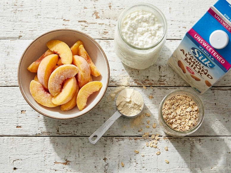 An overhead view of healthy peaches and cream smoothie ingredients arranged on a rustic white wooden table, featuring a bowl of frozen peach slices, cottage cheese, almond milk, vanilla protein powder, and rolled oats in bright, natural light.