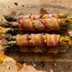 A bright overhead view of a delicious stack of golden-brown, crispy bacon wrapped asparagus bundles resting on a rustic wooden cutting board, scattered with coarse black pepper and sea salt.
