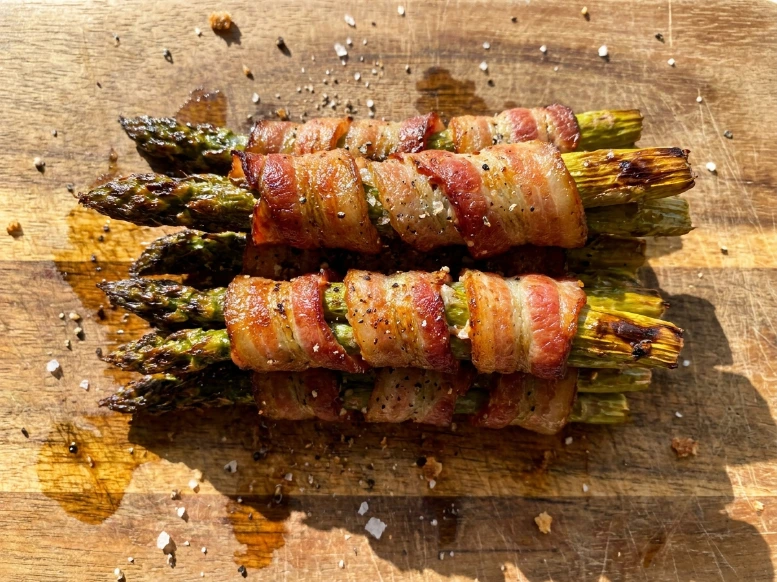 A bright overhead view of a delicious stack of golden-brown, crispy bacon wrapped asparagus bundles resting on a rustic wooden cutting board, scattered with coarse black pepper and sea salt.