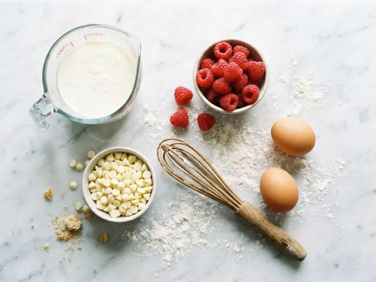 A baker in a linen apron using a wire whisk to mix flour and brown sugar in a large ceramic bowl to prepare the batter for raspberry white chocolate muffins.
