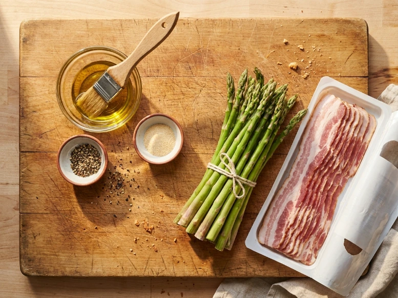 A bright overhead flat lay photograph on a scratched wooden cutting board showing the raw ingredients for the appetizer, including fresh asparagus spears, a package of standard-cut bacon, a bowl of olive oil, and small pinch bowls of spices.