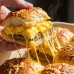 A candid, bright food photograph of a hand pulling a warm, gooey Crack Breakfast Slider away from a baking dish, showing melted cheddar cheese stretching from the savory sausage and fluffy eggs beneath a golden-brown bun.