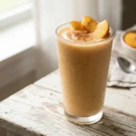 A tall glass filled with a thick, creamy peach smoothie garnished with fresh peach slices and a dusting of cinnamon. The drink sits on a weathered white table next to a sunny window, with a bowl of peaches in the soft background.