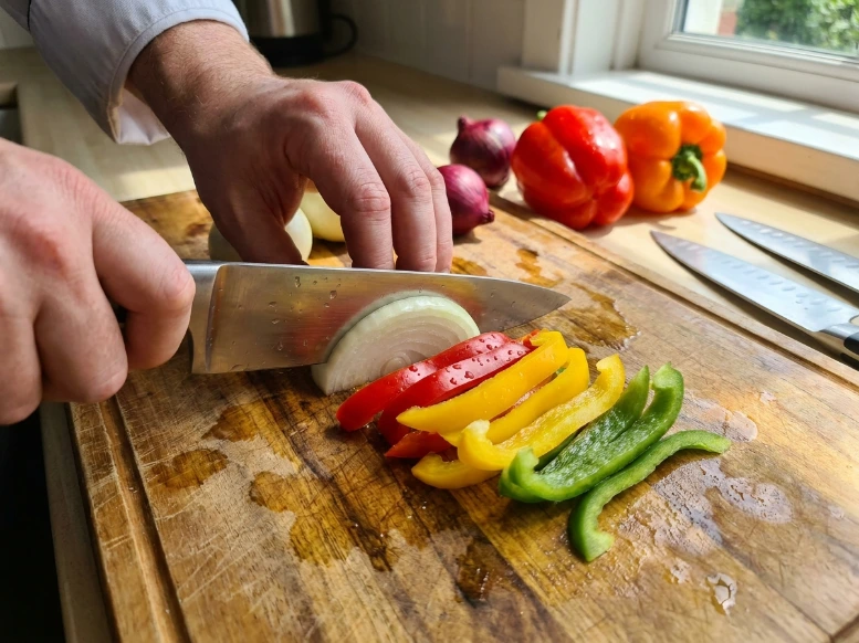 A candid, close-up shot of hands using a sharp knife to slice raw Italian sausage links into thick coins on a weathered wooden cutting board in a sunlit kitchen.