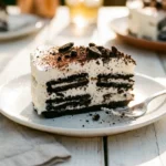 A close-up shot of a single slice of creamy, layered Oreo icebox cake served on a simple white plate, featuring distinct layers of chocolate cookies and whipped cream, topped with crushed cookie crumbs and cocoa powder.