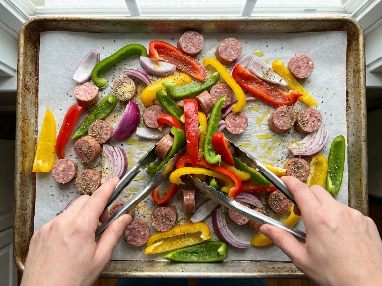 An overhead view of two hands using metal tongs to toss sliced sausage coins, bell peppers, and onions with olive oil and spices on a parchment-lined baking sheet.