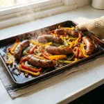 A bright, photorealistic shot of a rustic, dark baking sheet filled with sizzling, caramelized Italian sausages and vibrant roasted bell peppers and onions, held by a hand in a tan oven mitt on a sunny white kitchen counter.
