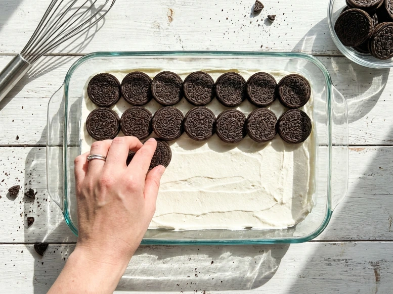 A person’s hand carefully arranging a single, neat layer of Oreo cookies over a base of whipped cream inside a glass baking dish to assemble a homemade Oreo icebox cake.