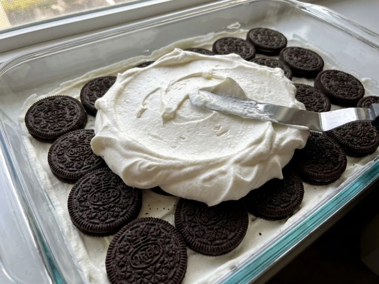 An offset spatula spreading a thick, fluffy layer of homemade whipped cream over a layer of Oreo cookies in a glass baking dish, illustrating the assembly process of a no-bake Oreo icebox cake.