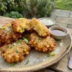A pile of crispy corn fritters with steam rising next to small bowls of sour cream and syrup on an outdoor patio.