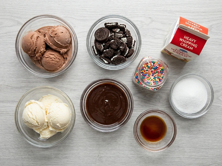 An overhead flat-lay shot of ingredients to make an ice cream cake at home, featuring glass bowls of chocolate and vanilla ice cream, hot fudge sauce, crushed Oreo cookies, sugar, and heavy cream.