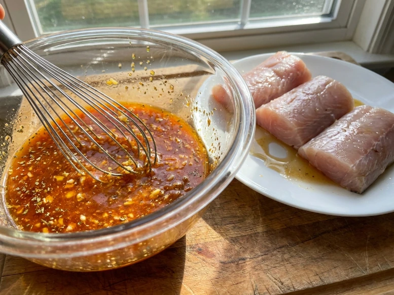 Whisking a savory garlic and lime mahi mahi marinade in a glass bowl next to raw fish fillets.