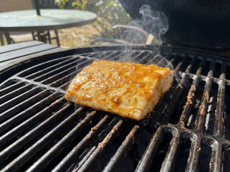 A single marinated fish fillet searing over high heat on a clean outdoor charcoal grill grate with light smoke.