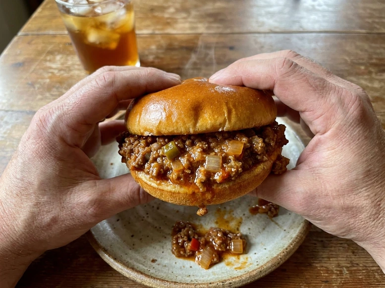 A first-person perspective shot showing a pair of hands holding a warm, juicy sloppy joe sandwich over a white ceramic plate as the thick sweet and tangy sauce drips down.