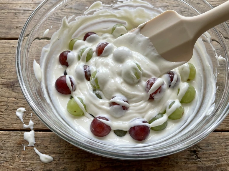 Folding whole seedless grapes into a thick white cream cheese and sour cream dressing mixture using a wide rubber spatula.