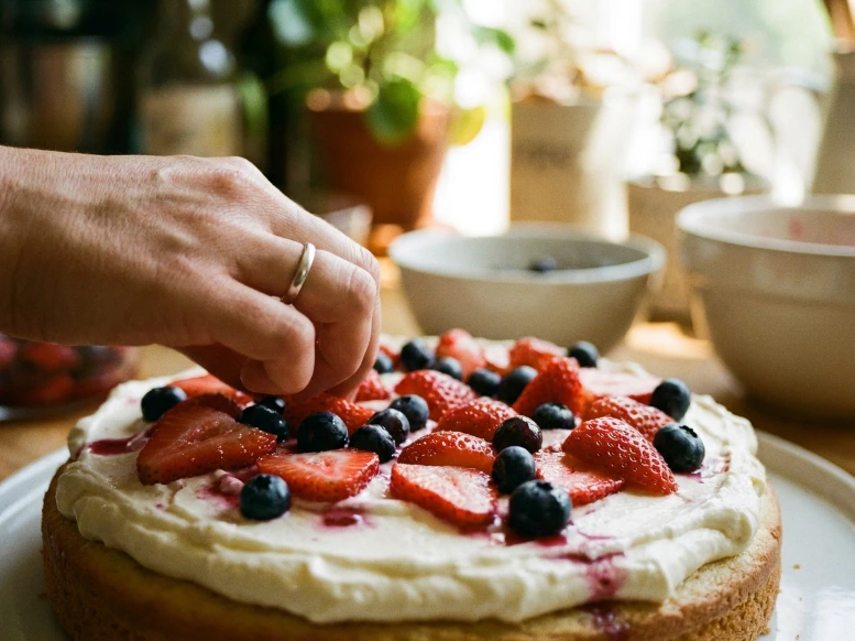 A candid shot of a baker's hand gently placing fresh sliced strawberries and blueberries onto a thick layer of mascarpone cake filling while assembling a homemade Berry Chantilly cake.