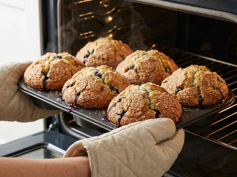 Hands wearing fabric oven mitts carefully pulling a hot metal tin of freshly baked jumbo blueberry muffins out of a steaming open oven.