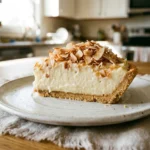 A perfect, tall slice of instant pudding coconut pie sitting on an elegant white ceramic plate over a rustic fringed linen cloth, topped with a thick layer of toasted coconut flakes.