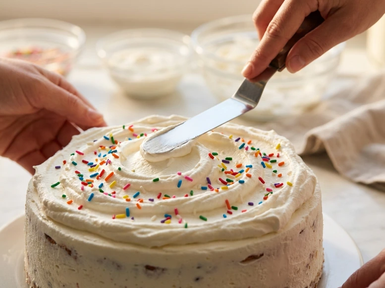 A close-up shot of an offset spatula creating decorative swirls in fresh whipped cream frosting on top of a round ice cream cake decorated with colorful sprinkles.